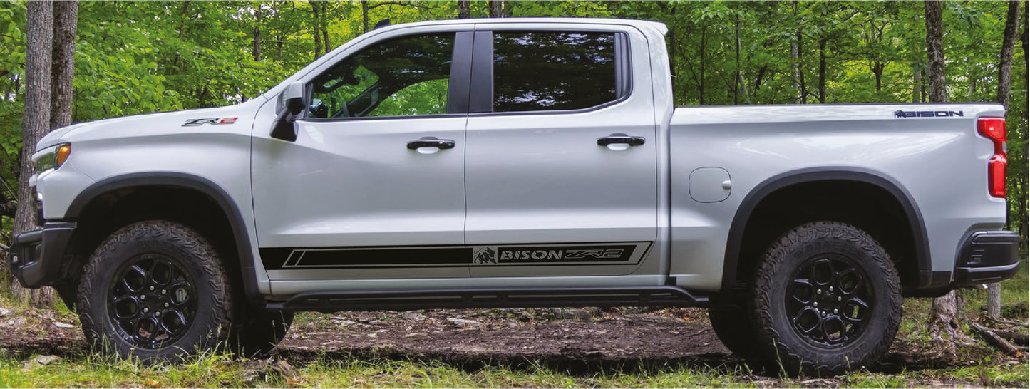 A white pickup truck with black wheels and black side decals reading 'BISON' is parked outdoors on a grassy path surrounded by green trees.